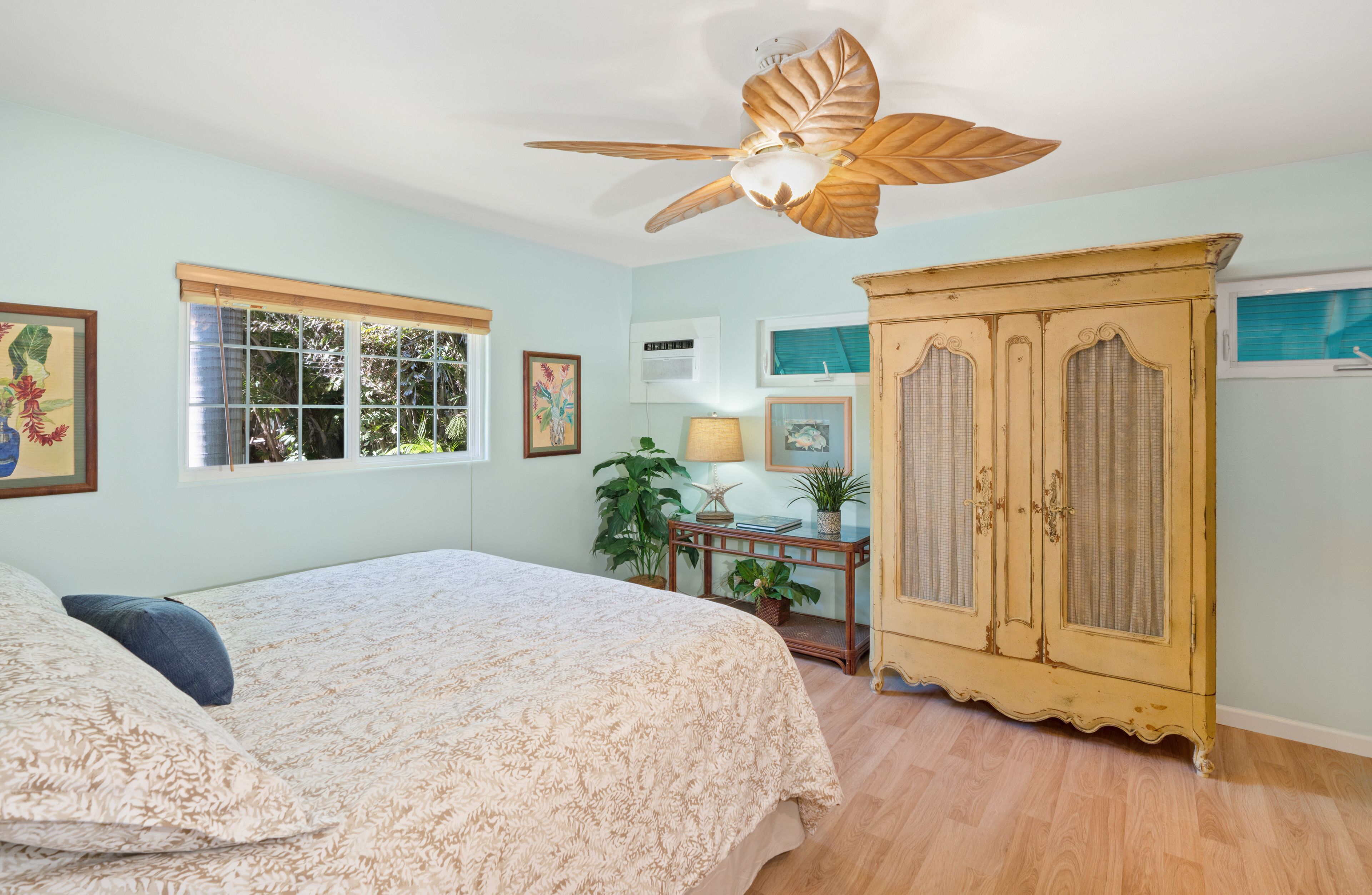 Un dormitorio con cama, ventilador de techo, un mueble de madera y ventana con vista a follaje.