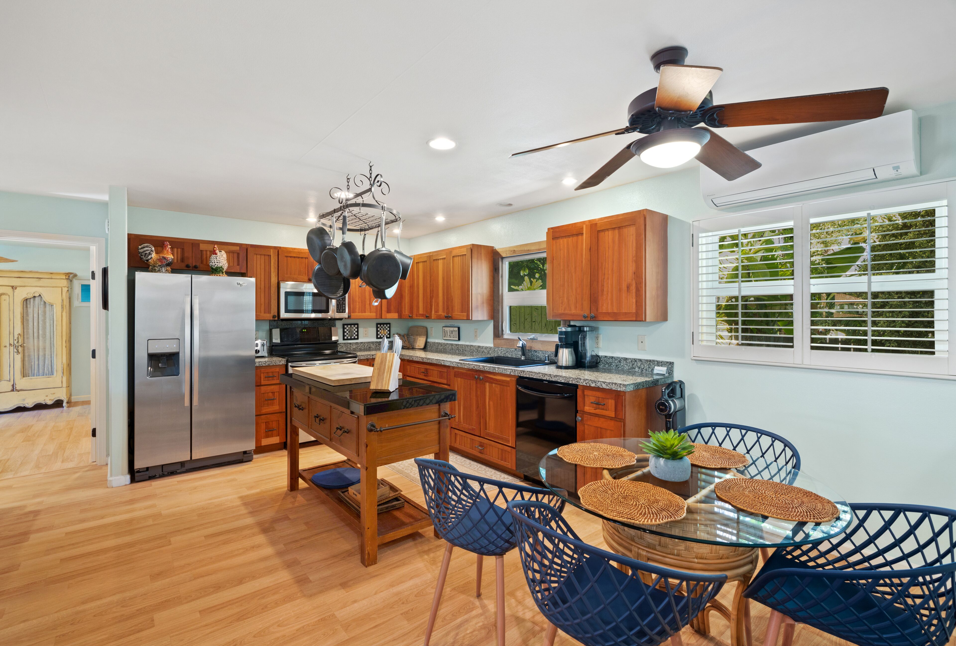 A modern kitchen with wooden cabinets, a stainless steel refrigerator, a dining area with blue chairs, and a ceiling fan.