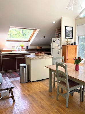 A compact kitchen with a small dining area, a trash bin, and a window with a view of greenery.