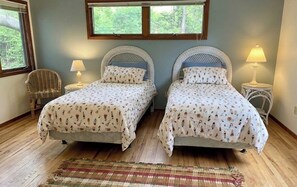 A room with two beds, a wicker chair, and a small table with a lamp.