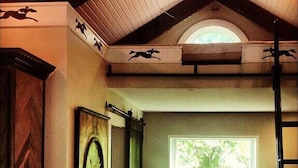 Une pièce avec un plafond en pente, une grande fenêtre donnant sur la verdure, une horloge au mur et une échelle appuyée contre le mur.