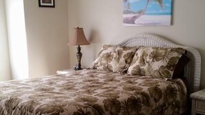 Une chambre à coucher avec un lit, un ventilateur de plafond, une table de chevet, une lampe et un tableau représentant une scène de plage.