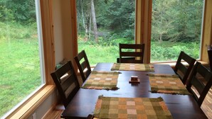A dining table with four chairs, a placemat, and a view of a forest through large windows. - Unique Home in Willard, Wa. 40 acres, 50 feet from Little White Salmon River (Willard)