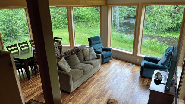 A spacious living area with a wooden floor, a sofa, two armchairs, and a dining table with chairs. Large windows offer a view of a lush garden and a river. - Unique Home in Willard, Wa. 40 acres, 50 feet from Little White Salmon River (Willard)