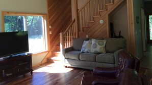 A living room with a wooden staircase, a sofa, a leather armchair, and a flat-screen TV on a wooden cabinet. - Unique Home in Willard, Wa. 40 acres, 50 feet from Little White Salmon River (Willard)