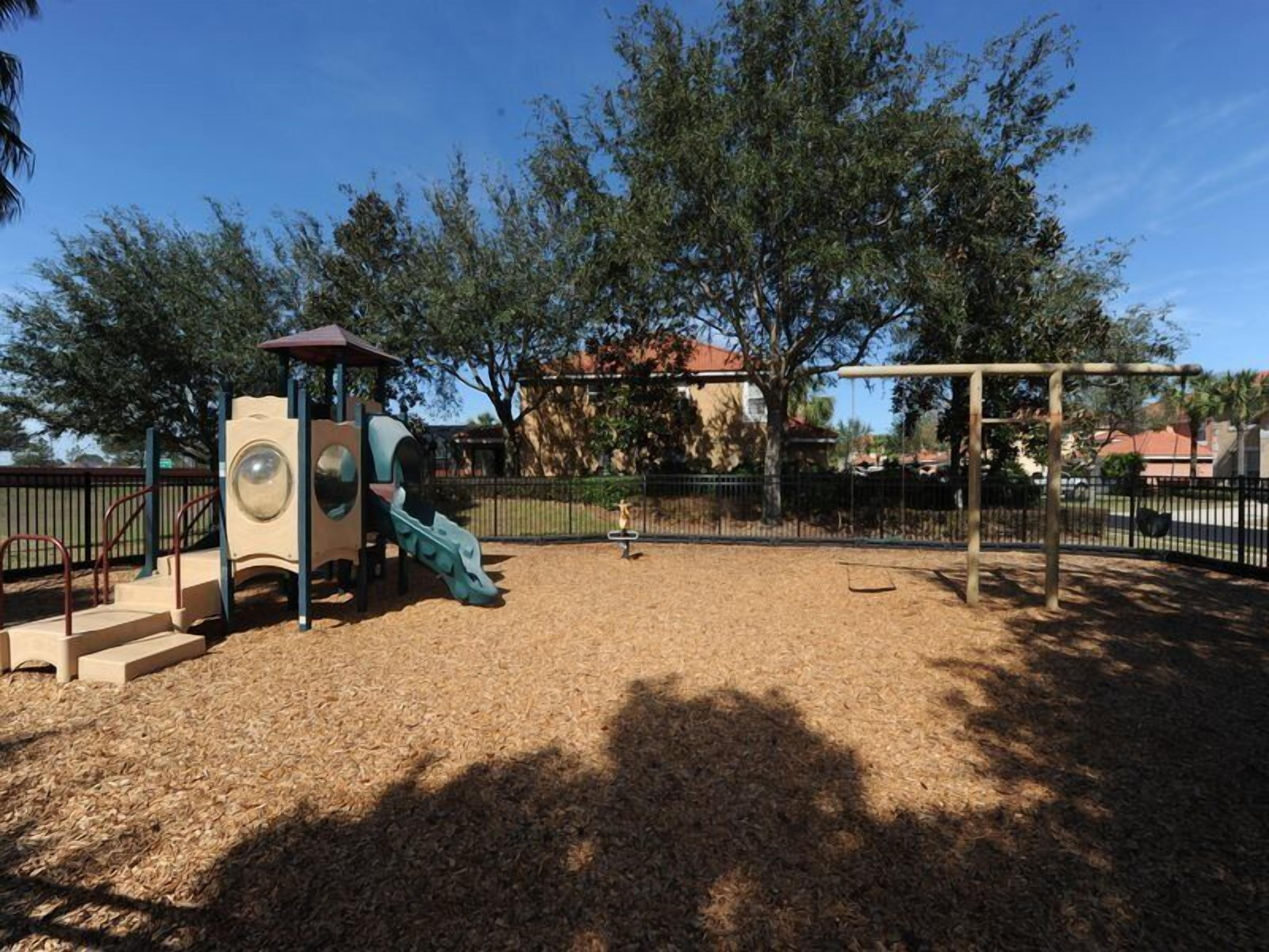 A playground with a slide and climbing structures, surrounded by trees and a fence.