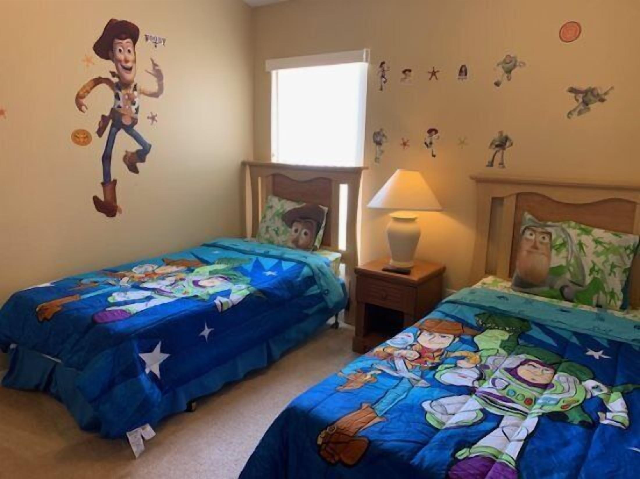 A bedroom with two beds, each covered with a blue blanket featuring cartoon characters. There is a wall decal of a cowboy character, a window, and a nightstand with a lamp.