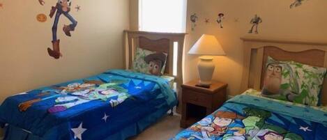 A bedroom with two beds, each covered with a blue blanket featuring cartoon characters. There is a wall decal of a cowboy character, a window, and a nightstand with a lamp.