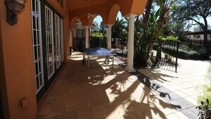 Eine überdachte Terrasse mit Tischtennisplatte, Deckenventilator und Blick auf einen Garten mit Palmen.