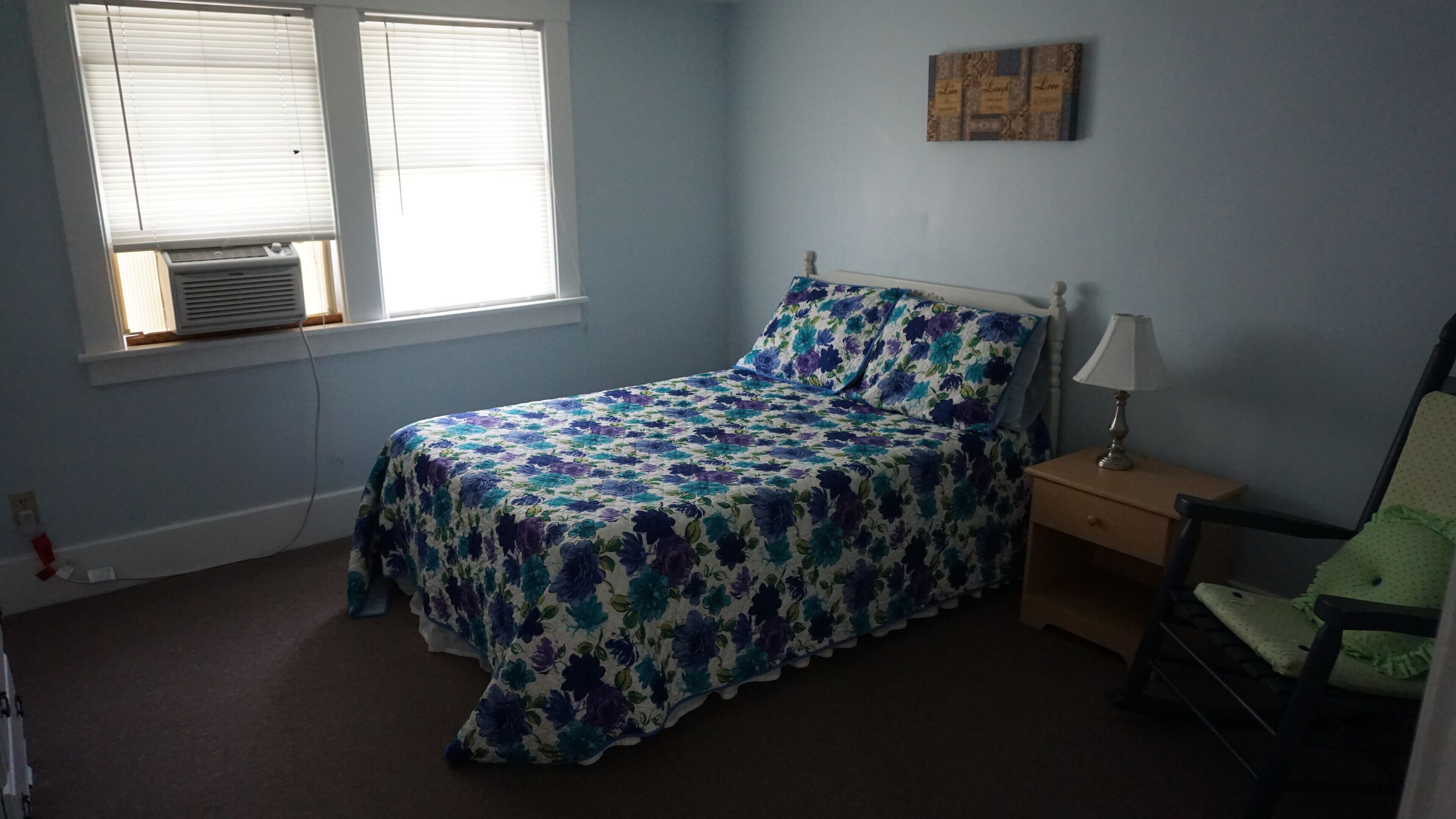 A bedroom with a bed, a nightstand, a chair, and a window with blinds.