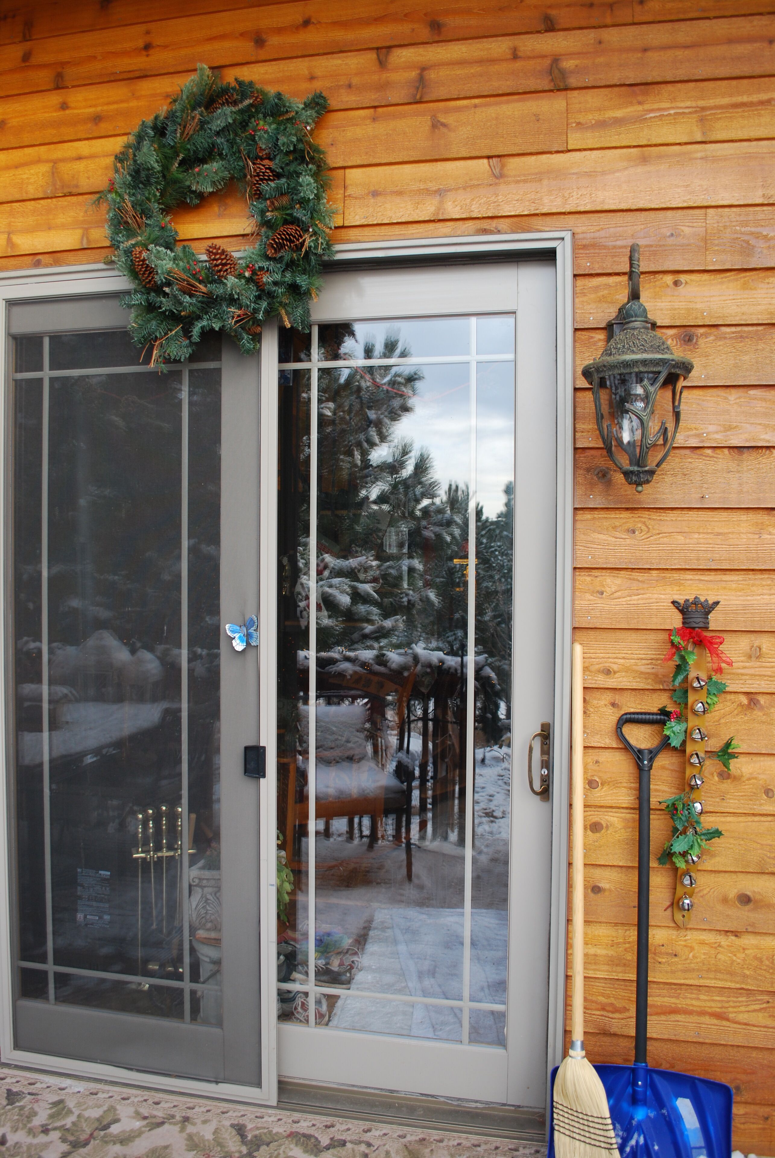 Une cabane en bois avec une porte en verre, une couronne de Noël, une lanterne et un balai appuyé contre le mur.