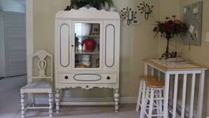 Una habitación con un armario blanco de puertas de cristal, una silla de madera, una mesita con taburetes y un reloj fijado a la pared.
