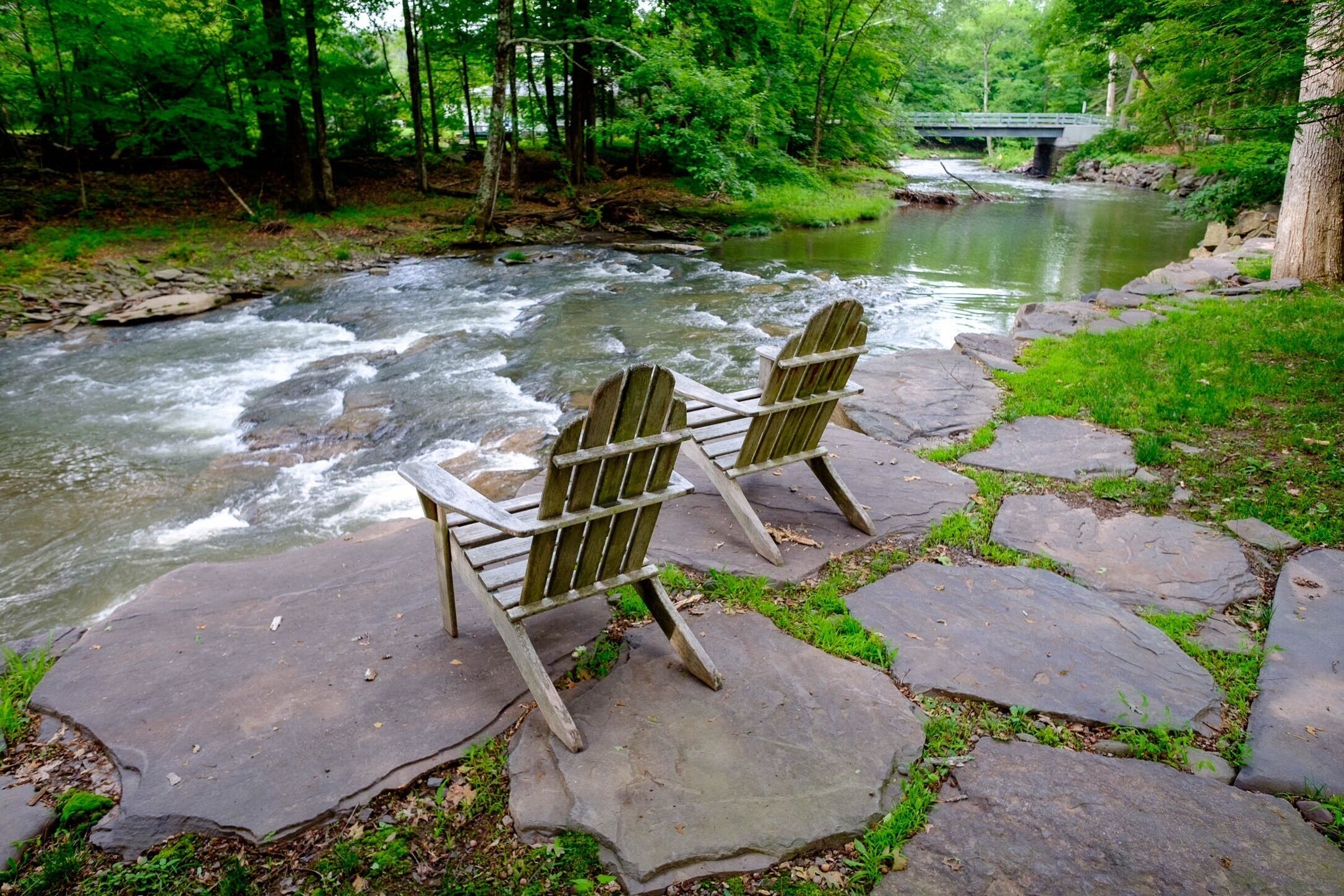 Dos sillas Adirondack mirando un río con un puente al fondo.