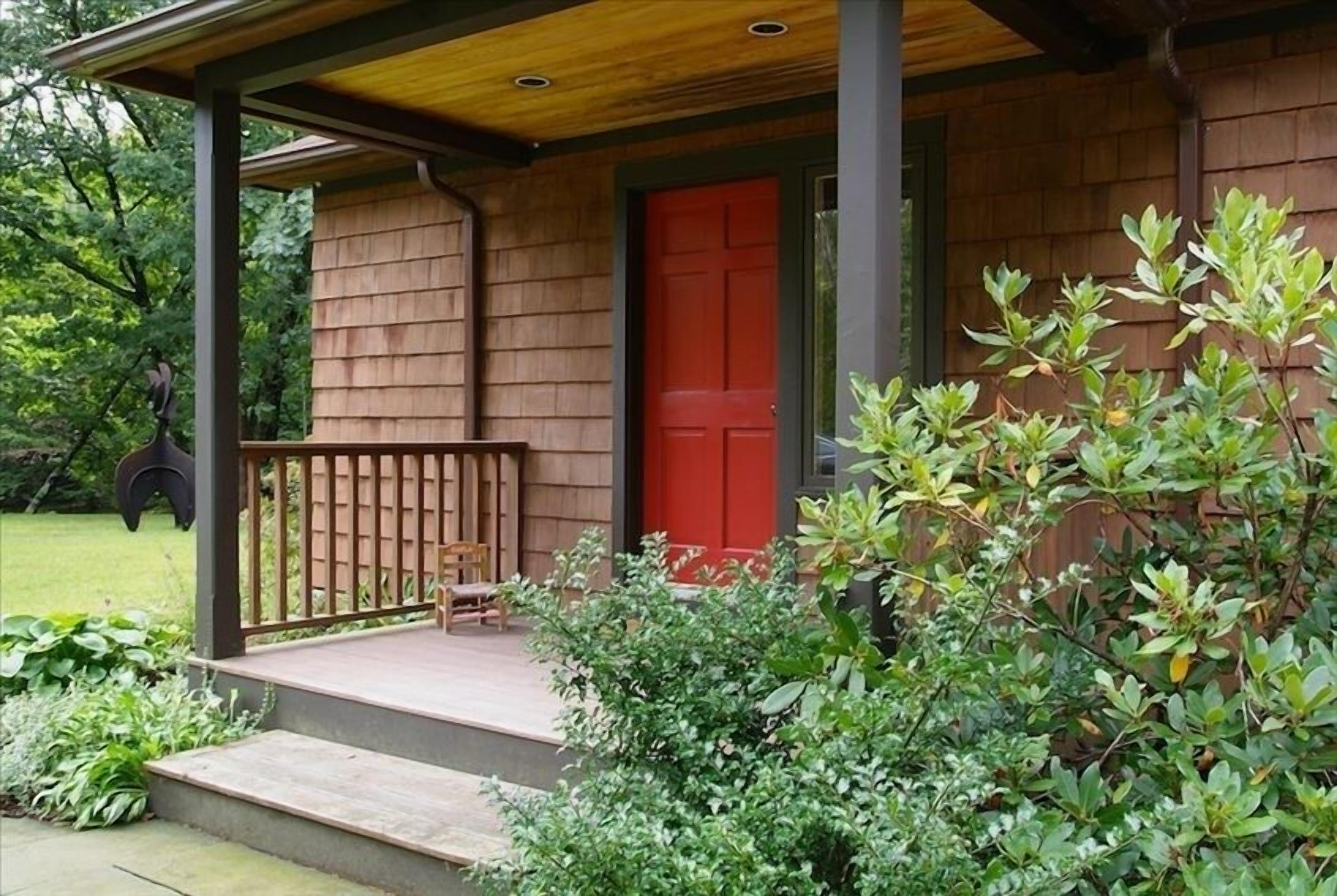 Une véranda en bois avec une porte rouge, des marches menant à l’entrée et un petit espace de repos avec un banc et une chaise.