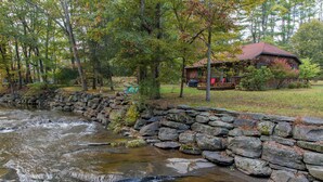 A stone wall beside a flowing river, surrounded by trees and a wooden cabin. - Streamside Cottage Half Mile to Town of Woodstock (Woodstock)