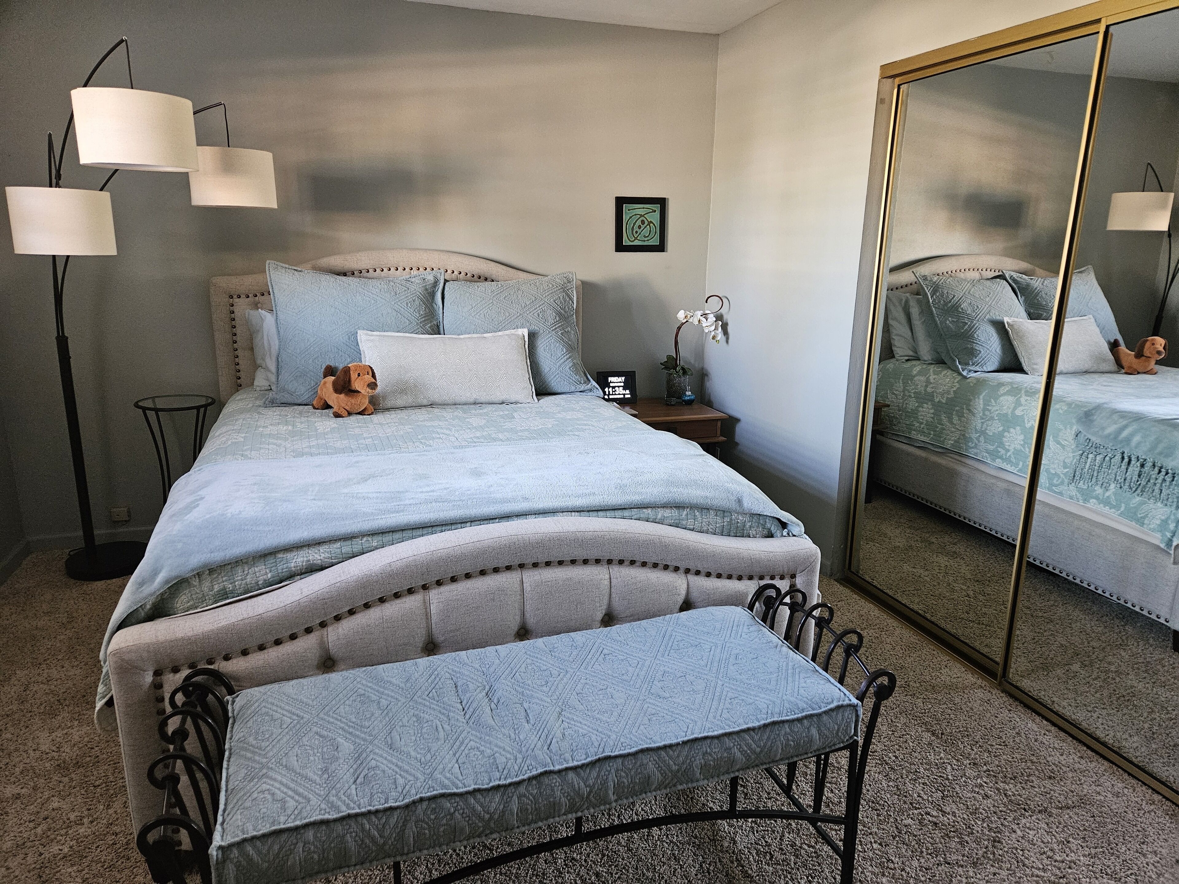 A neatly arranged bedroom with a large bed, a bedside table, a mirror, and a floor lamp.