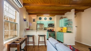 Une cuisine compacte avec des meubles en bois, un réfrigérateur, un four à micro-ondes et une cuisinière. On y trouve deux tabourets et un canapé dans l’espace de vie adjacent.