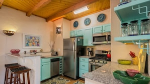 Une cuisine avec des armoires de couleur turquoise, un réfrigérateur en acier inoxydable, un four à micro-ondes et une cuisinière. On y trouve des tabourets au comptoir, un évier et un tapis à motifs sur le sol.