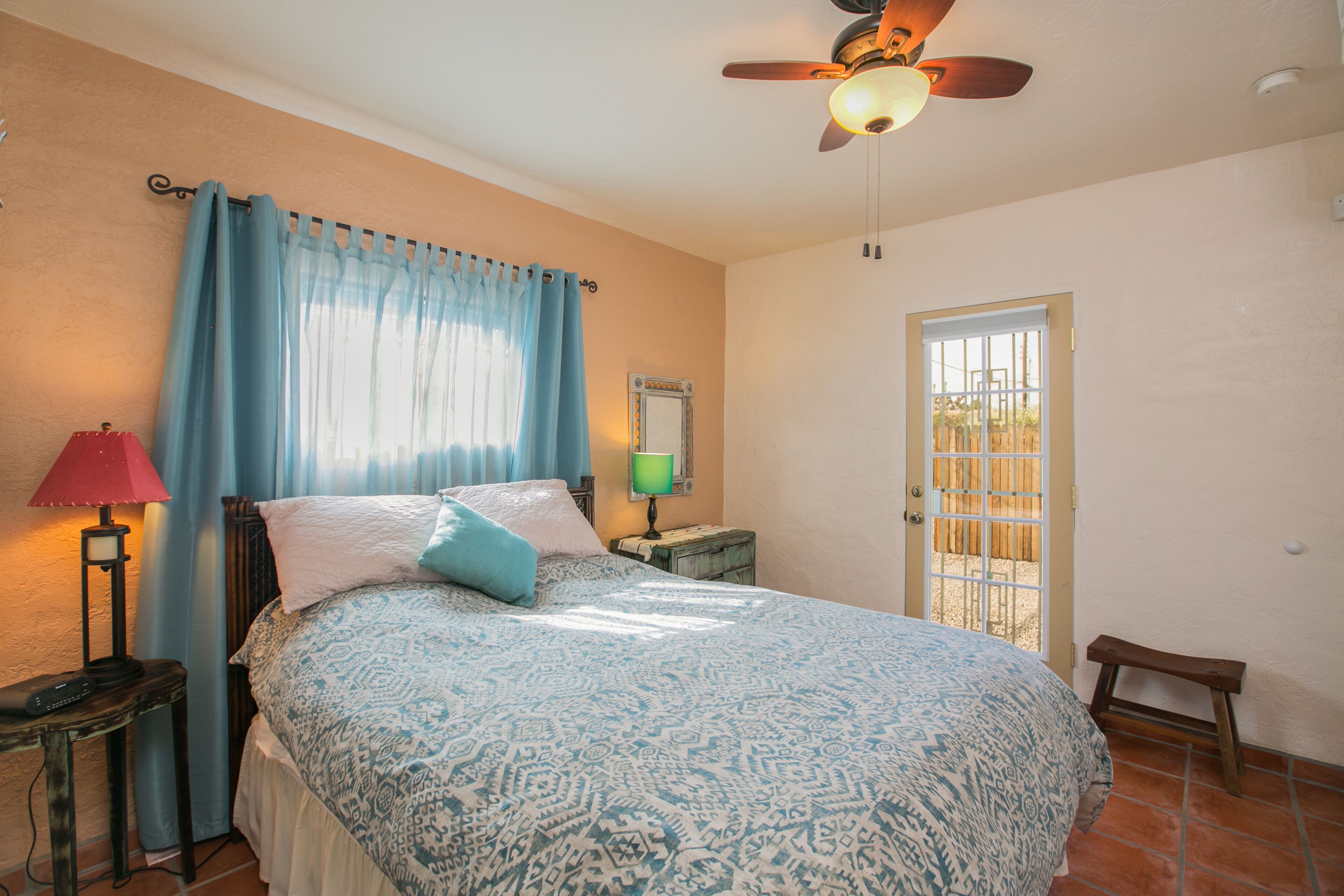 A bedroom with a bed, a nightstand, a lamp, a ceiling fan, a window with curtains, and a door.