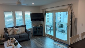 A living room with a grey sofa, a coffee table, and a TV on the wall. There are windows with blinds, a balcony with a railing, and a ceiling fan. - SEPTEMBER BEACH TIME... NO CROWDS..LOW PRICES! (Gulf Shores)