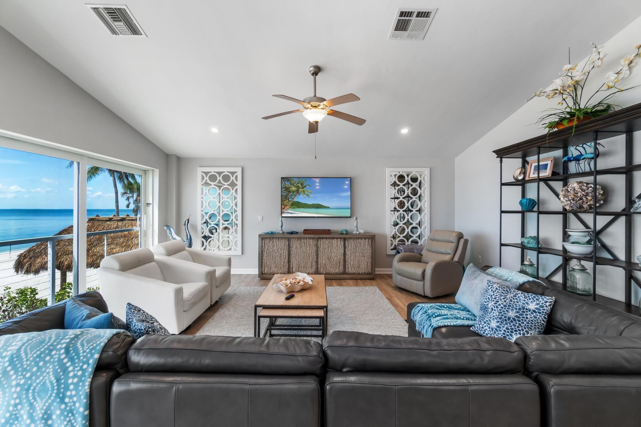 A spacious living room with a large TV, comfortable seating, a ceiling fan, and a view of the ocean.