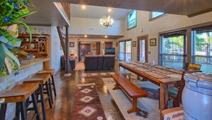 A spacious living area with a high ceiling, a chandelier, a long wooden table with benches, and a large window providing natural light.