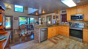 A spacious kitchen with wooden cabinets, a built-in dishwasher, a microwave, and an oven. There is a large island with a sink and a chair. The kitchen has a stone floor and a view of the outdoors through large windows.