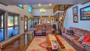 A spacious living room with a large brown leather sofa, a wooden coffee table, and a patterned rug. There are multiple windows with blinds, a chandelier, and framed artwork on the walls.