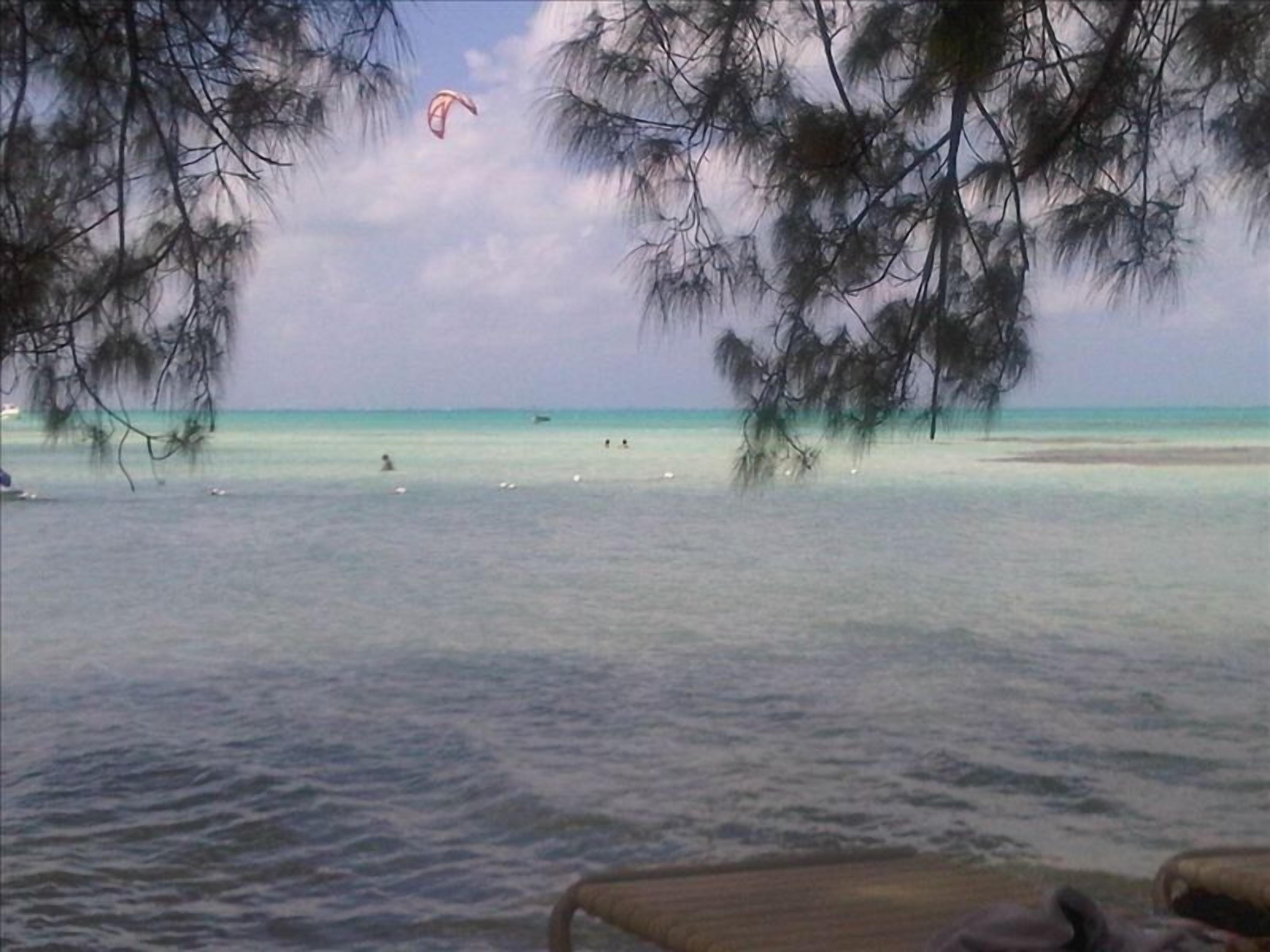 Een strand met helder turquoise water, mensen die zwemmen en een paraschuttersboot in de verte.