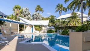 A swimming pool with lounge chairs and a covered seating area, surrounded by palm trees and a building with a white roof.