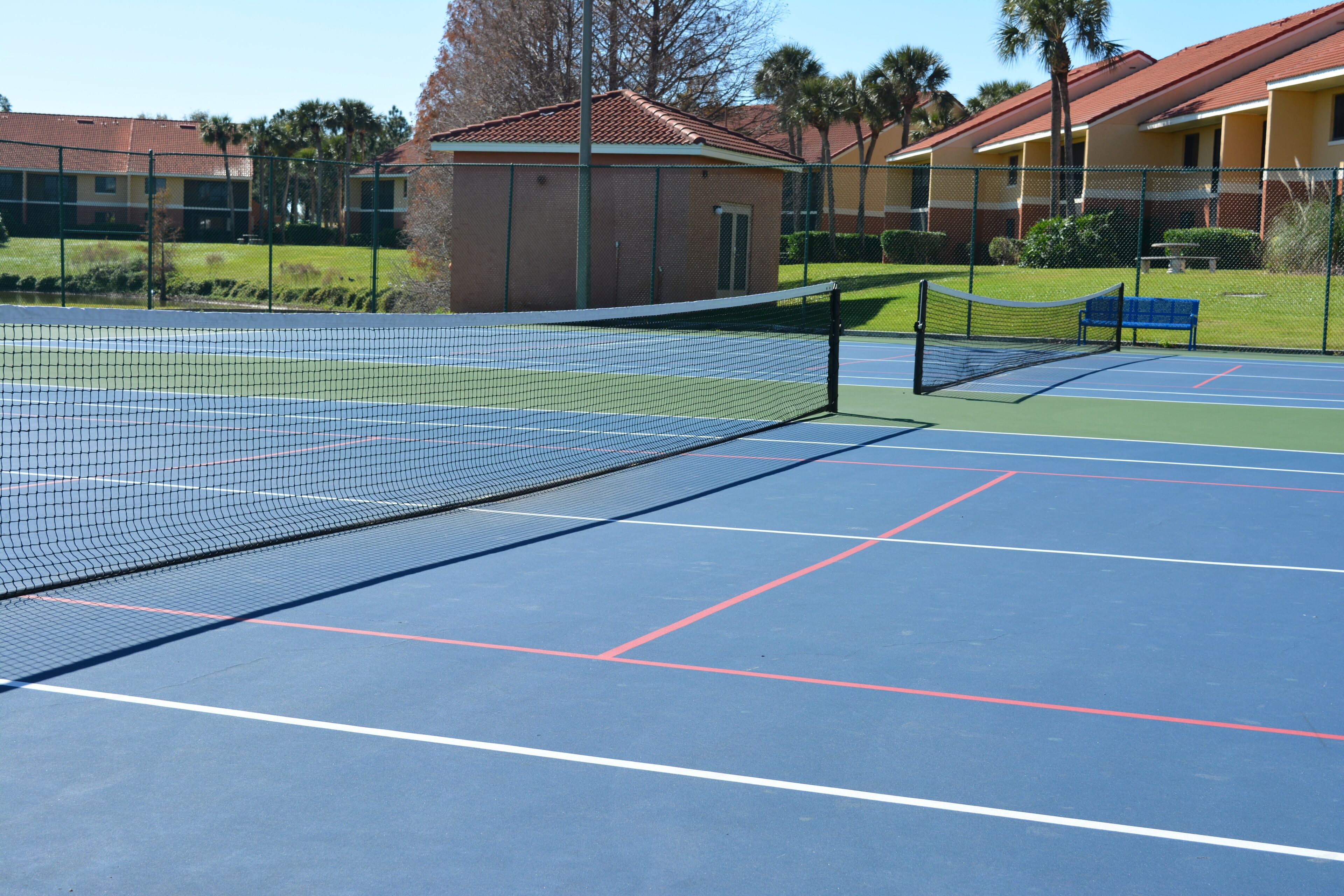 Ein Tennisplatz mit Netz, umzäunt und mit einem Gebäude mit rotem Dach.