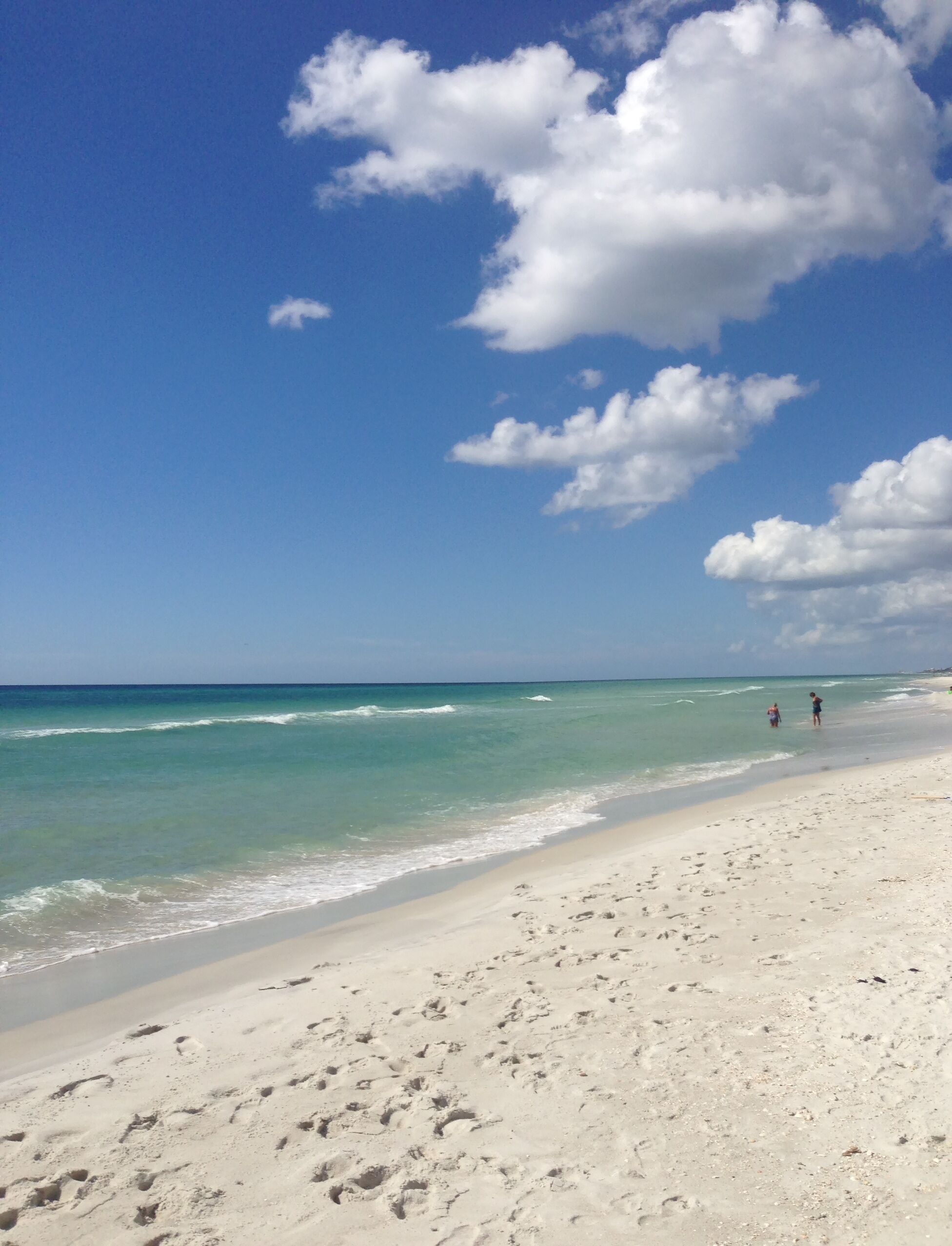 A beach with clear turquoise water, white sand, and a few people walking along the shore.