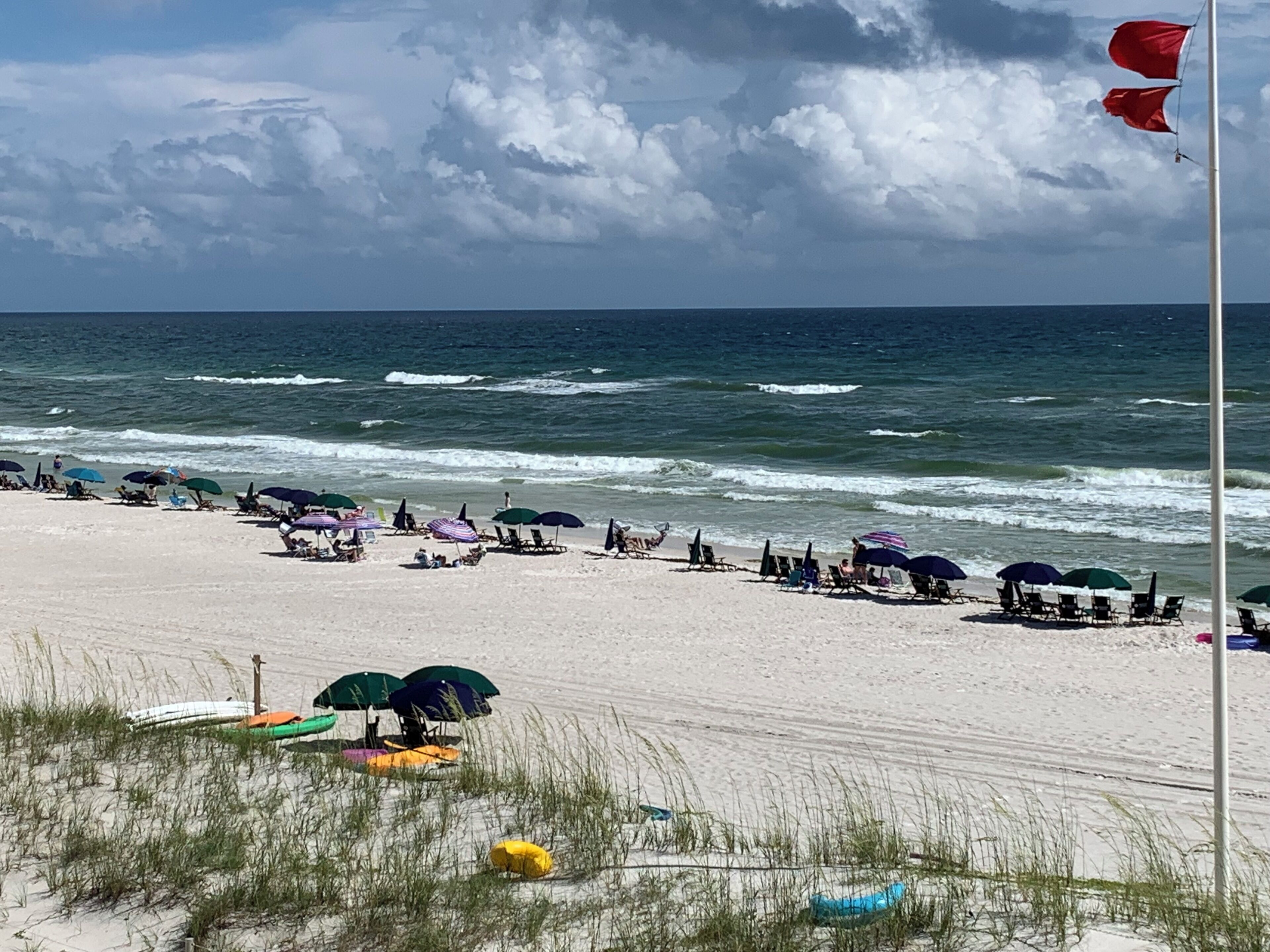 Une plage avec des parasols et des chaises, un mât de drapeau et l’océan en arrière-plan.