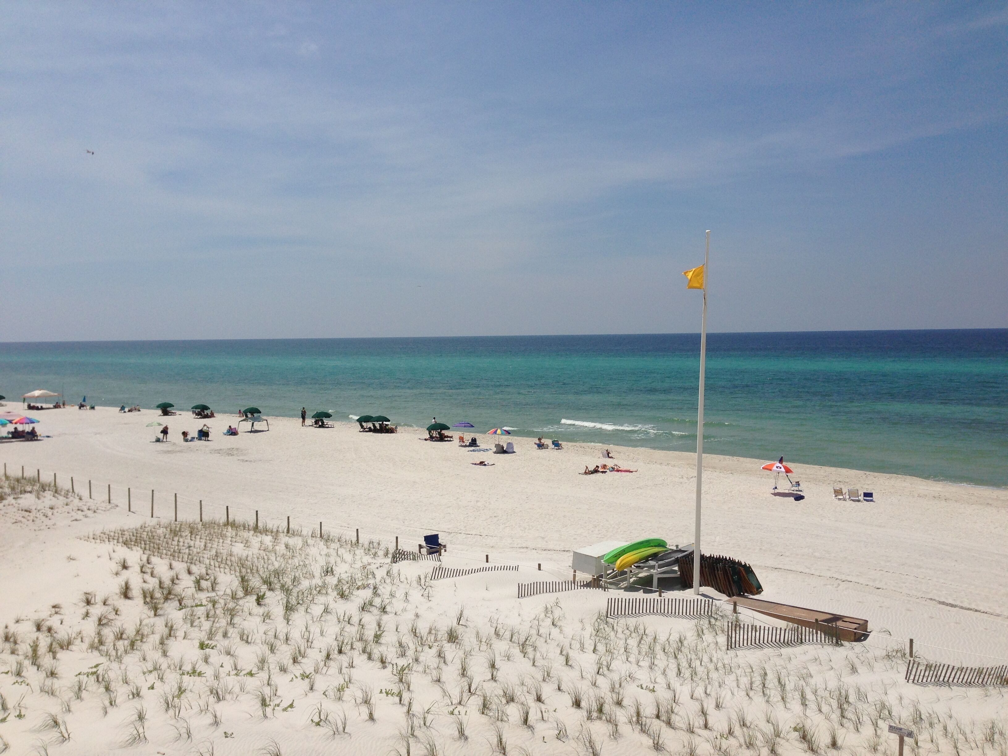 A sandy beach with umbrellas and beach chairs, a flagpole with a flag, and a lifeguard tower.