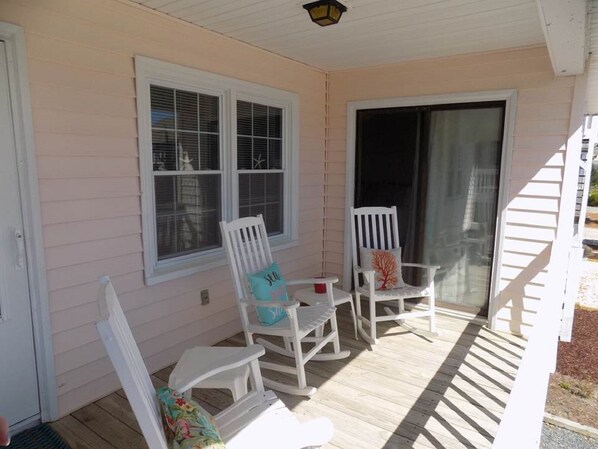 A covered porch with white rocking chairs and a small table, featuring a sliding glass door and windows. - Wonderful location, mid-island! Short walk to beach, pier and island market. (Sunset Beach)