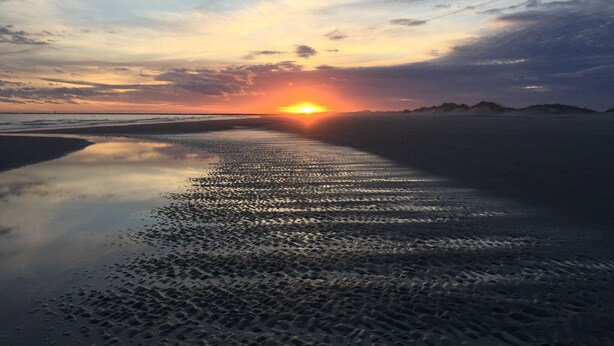 Een strandscène met een zonsondergang boven het water, een zandstrand en heuvels in de verte.