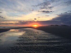 En strandscen med en solnedgång över vattnet, en sandstrand och avlägsna kullar.