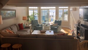 Un salon comprenant un canapé, des fauteuils, une table basse et un téléviseur. Une fenêtre offre une vue sur la mer.