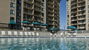 Una piscina dalle acque cristalline, circondata da lettini e ombrelloni, adiacente a un grattacielo con balconi.