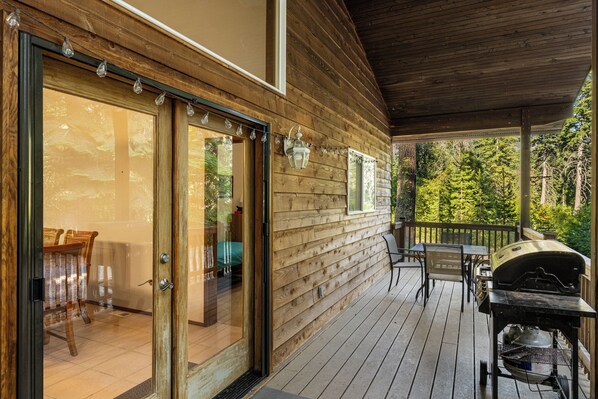 Een houten terras met een glazen deur naar een kamer, tuinmeubilair en een barbecue.