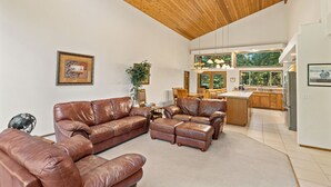 A spacious living area with a high wooden ceiling, a brown leather sofa set, a dining area with wooden chairs, and a kitchen with large windows. - Elk Tracks Lodge (Easton)