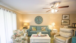A living room with a ceiling fan, a clock, and a fish-themed wall decoration. - Happy Place Chateau La Mer II #1301 2-Bd 2-Bth Ground Floor Private Patio (Destin)