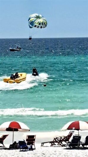 A beach with a parasail, a speedboat, and a rowboat. There are beach chairs and umbrellas.