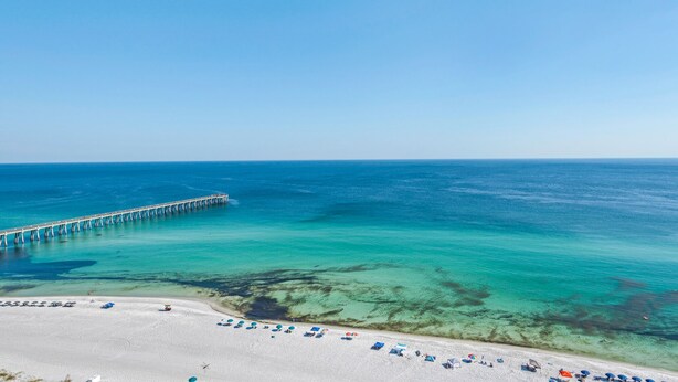 Une plage avec une jetée qui s’avance dans l’océan, une eau turquoise et limpide, et une plage de sable fin parsemée de parasols et de chaises longues.