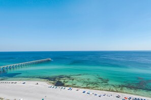 Una playa con un muelle que se adentra en el océano, aguas turquesas cristalinas y una playa de arena con sombrillas y tumbonas.