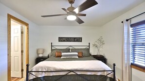 Ein Schlafzimmer mit Bett, Deckenventilator, Nachttisch, Lampe, Fenster mit Jalousien und einer Tür.