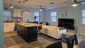 A modern kitchen with a central island, a dining area with a TV, and a living room with a sofa and chairs.