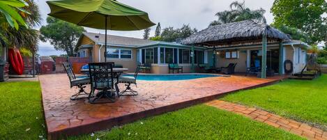 A backyard with a pool, a covered patio, and a thatched-roof structure.