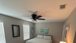 Une chambre avec deux lits, un ventilateur de plafond, une table de chevet avec une lampe et une étagère fixée au mur.