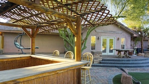 Un espace bar extérieur doté d’une pergola en bois, de sièges et d’un chemin en pierre menant à une maison.